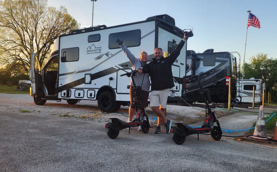 Kenny and Brittnay cheering with their arms in the air next to their scooters and their Jayco Granite Ridge RV at sunrise