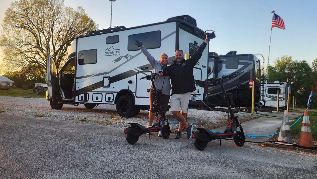 Kenny and Brittnay cheering with their arms in the air next to their scooters and their Jayco Granite Ridge RV at sunrise